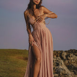 A woman stands barefoot on grass near rocks, wearing the Ananda Lālita Dress Blush Rose – Crinkle Linen Goddess Dress by AYA Sacred Wear. Her long, wavy hair with flowers and brass rings frames her soft gaze beneath a purple-blue sky.