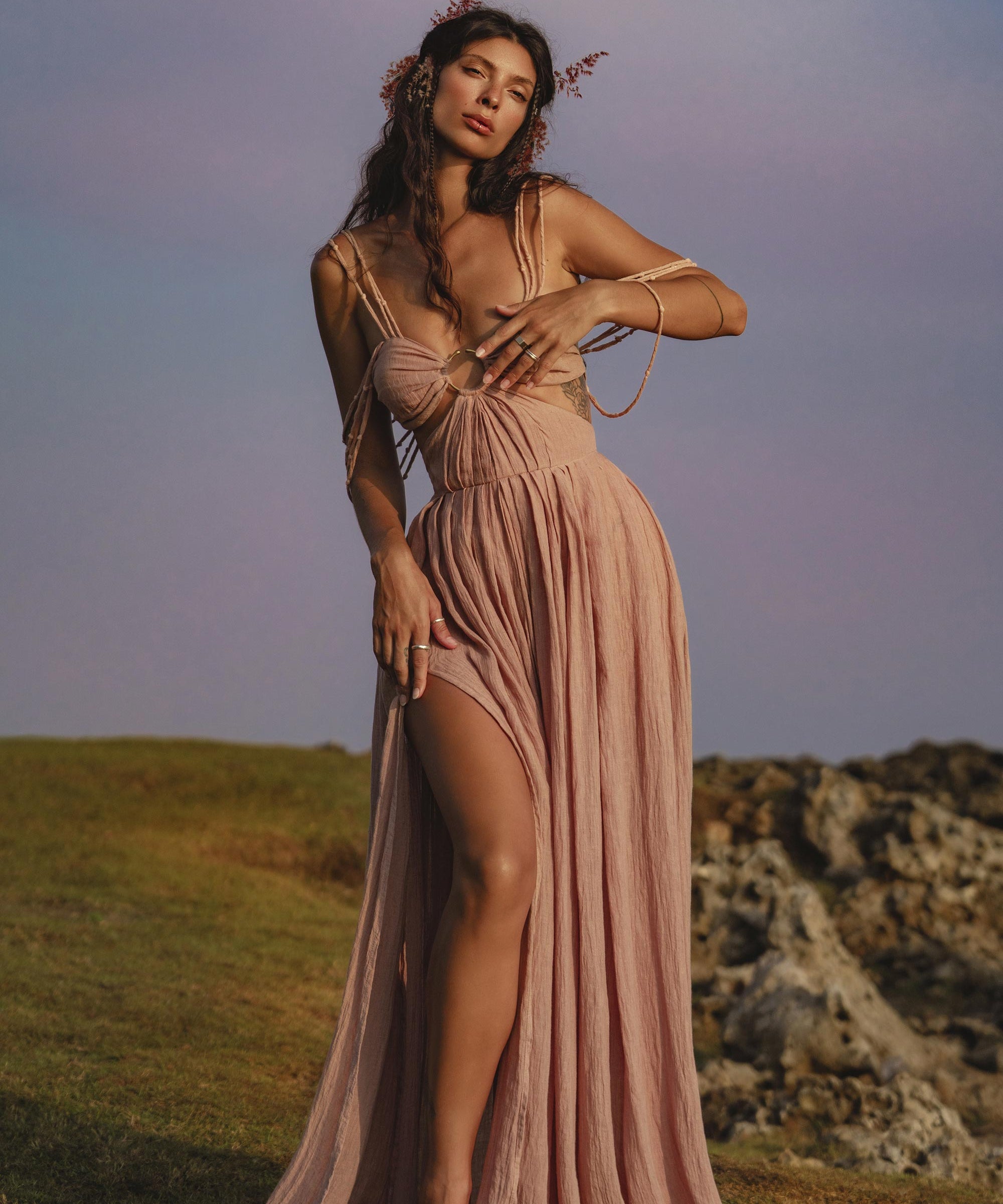 A woman stands barefoot on grass near rocks, wearing the Ananda Lālita Dress Blush Rose – Crinkle Linen Goddess Dress by AYA Sacred Wear. Her long, wavy hair with flowers and brass rings frames her soft gaze beneath a purple-blue sky.