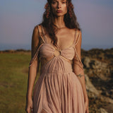 A woman stands on grassy terrain near rocks, wearing the Ananda Lālita Dress Blush Rose – Crinkle Linen Goddess Dress by AYA Sacred Wear. Her long brown hair shines in the sun, adorned with flowers and handcrafted brass rings under a pastel sky.