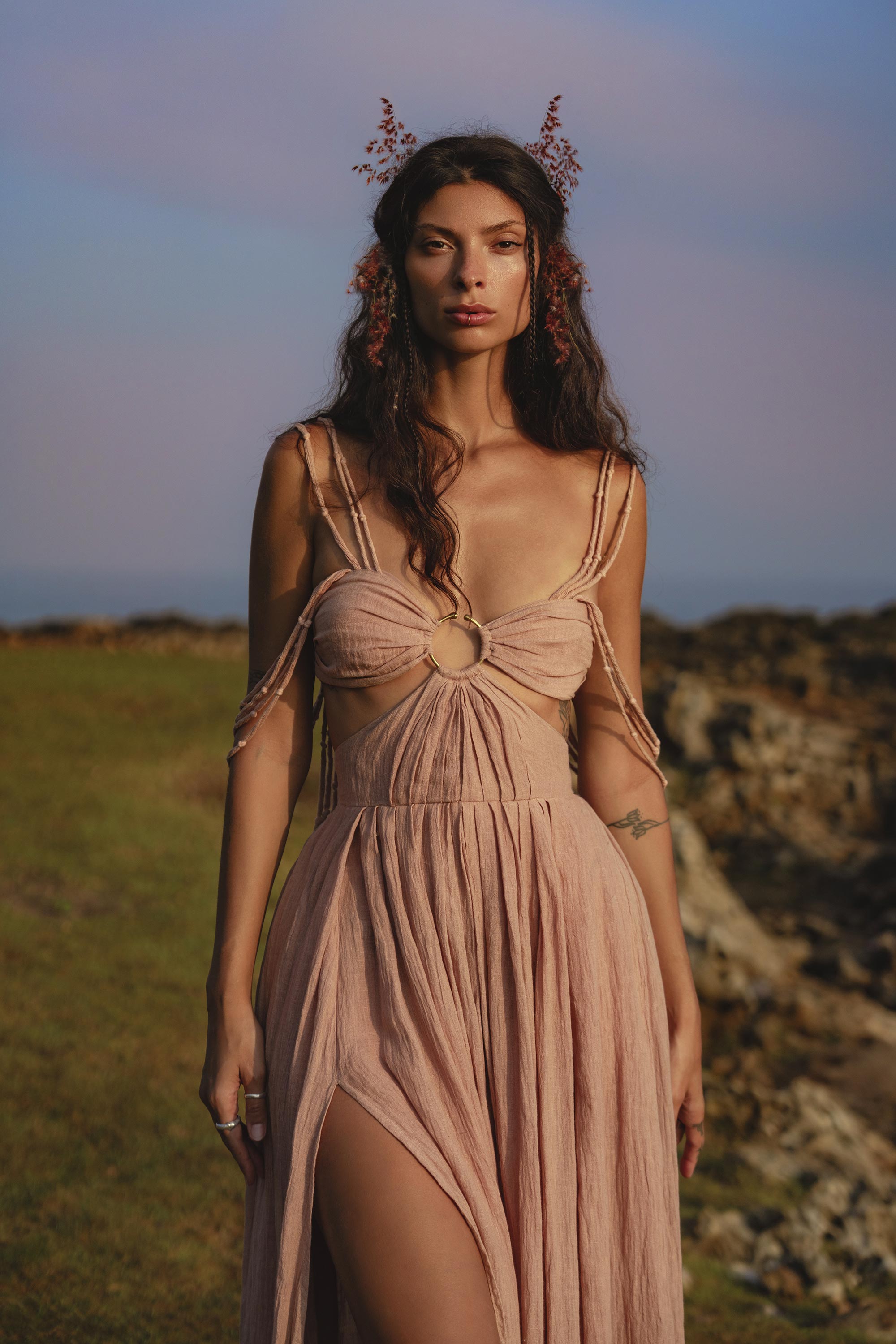A woman stands on grassy terrain near rocks, wearing the Ananda Lālita Dress Blush Rose – Crinkle Linen Goddess Dress by AYA Sacred Wear. Her long brown hair shines in the sun, adorned with flowers and handcrafted brass rings under a pastel sky.
