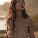 A woman with long wavy brown hair and small pink flowers gazes upward, wearing the Ananda Lālita Dress Blush Rose – Crinkle Linen Goddess Dress by AYA Sacred Wear. Soft sunlight and floating seeds create a dreamy outdoor scene.