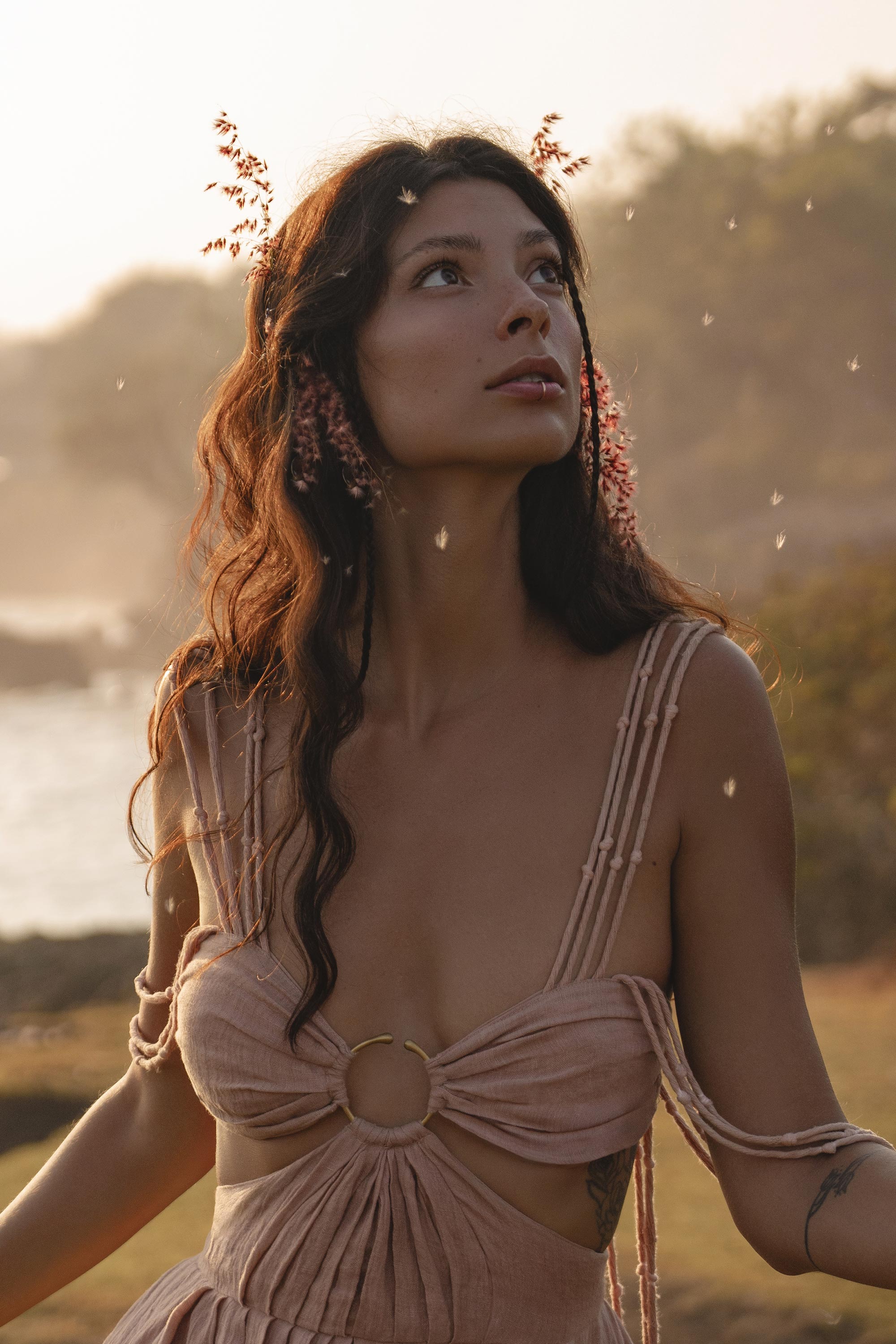 A woman with long wavy brown hair and small pink flowers gazes upward, wearing the Ananda Lālita Dress Blush Rose – Crinkle Linen Goddess Dress by AYA Sacred Wear. Soft sunlight and floating seeds create a dreamy outdoor scene.