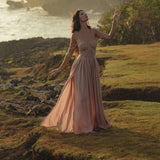 A woman in the Ananda Lālita Dress Blush Rose – Crinkle Linen Goddess Dress by AYA Sacred Wear stands on grassy, rocky terrain by the sea at sunset, rings on her fingers, golden light around her, and lush foliage and waves in the background.