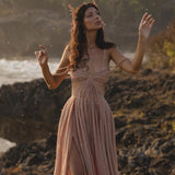 A woman wears the Ananda Lālita Dress Blush Rose – Crinkle Linen Goddess Dress by AYA Sacred Wear, standing on rocky terrain near water, arms raised beneath falling particles and soft sunlight, exuding an ethereal, mystical aura.