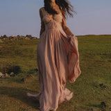 A woman with long dark hair strolls on grass beneath a blue sky, wearing the Ananda Lālita Dress Blush Rose – Crinkle Linen Goddess Dress by AYA Sacred Wear. She holds her flowing dress and wears floral headpieces for an ethereal look.
