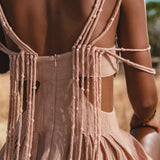 A close-up shows a woman’s back in the Ananda Lālita Dress Blush Rose – Crinkle Linen Goddess Dress by AYA Sacred Wear, featuring knotted draped strings and handcrafted brass rings, glowing in sunlight amid dry grass and greenery.