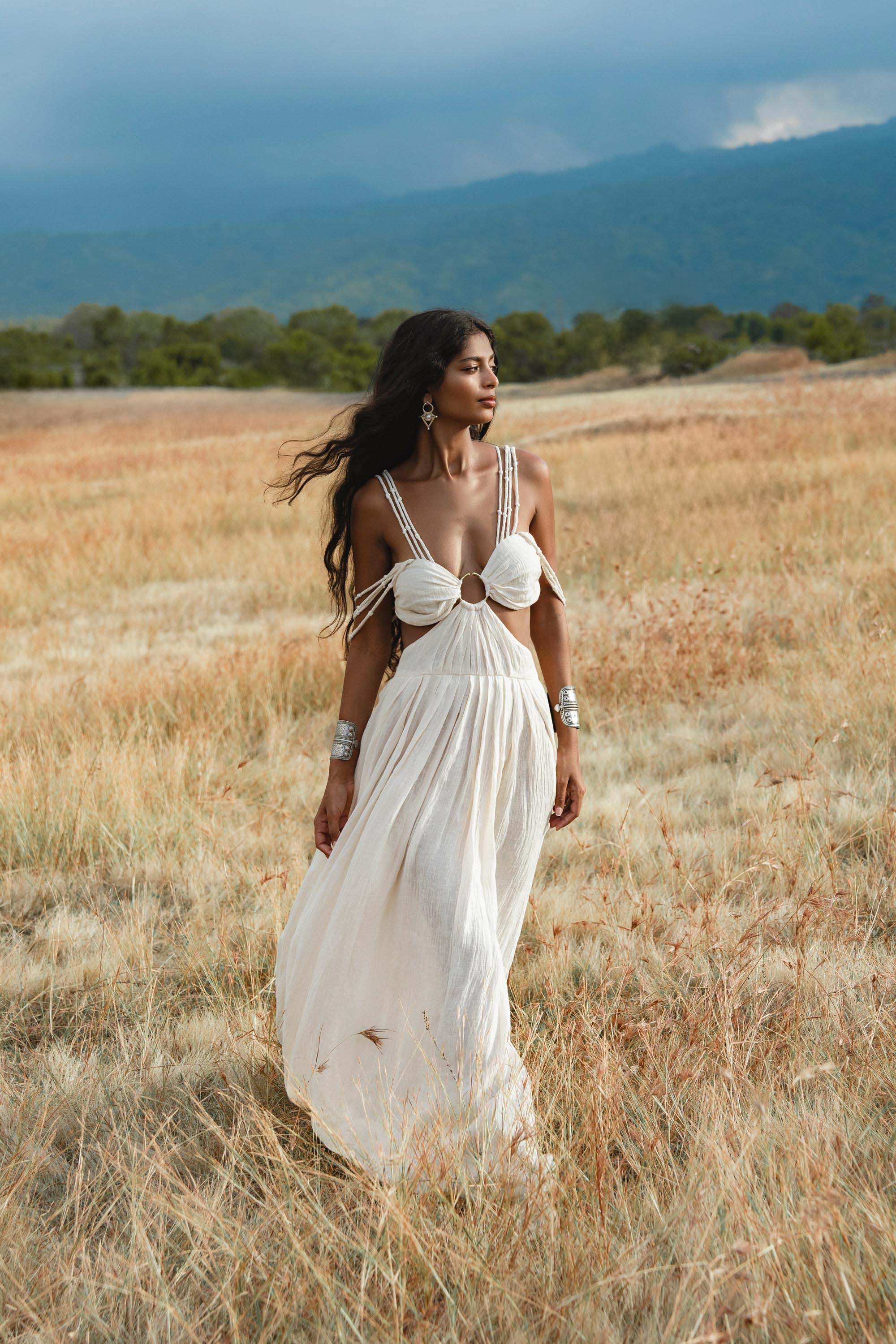 A woman in the Ananda Lālita Dress Ivory Colour – Linen Organic Goddess Gown by AYA Sacred Wear strolls through a sunlit grassy field, her long dark hair flowing and handcrafted brass rings gleaming, with mountains and clouds in the background.