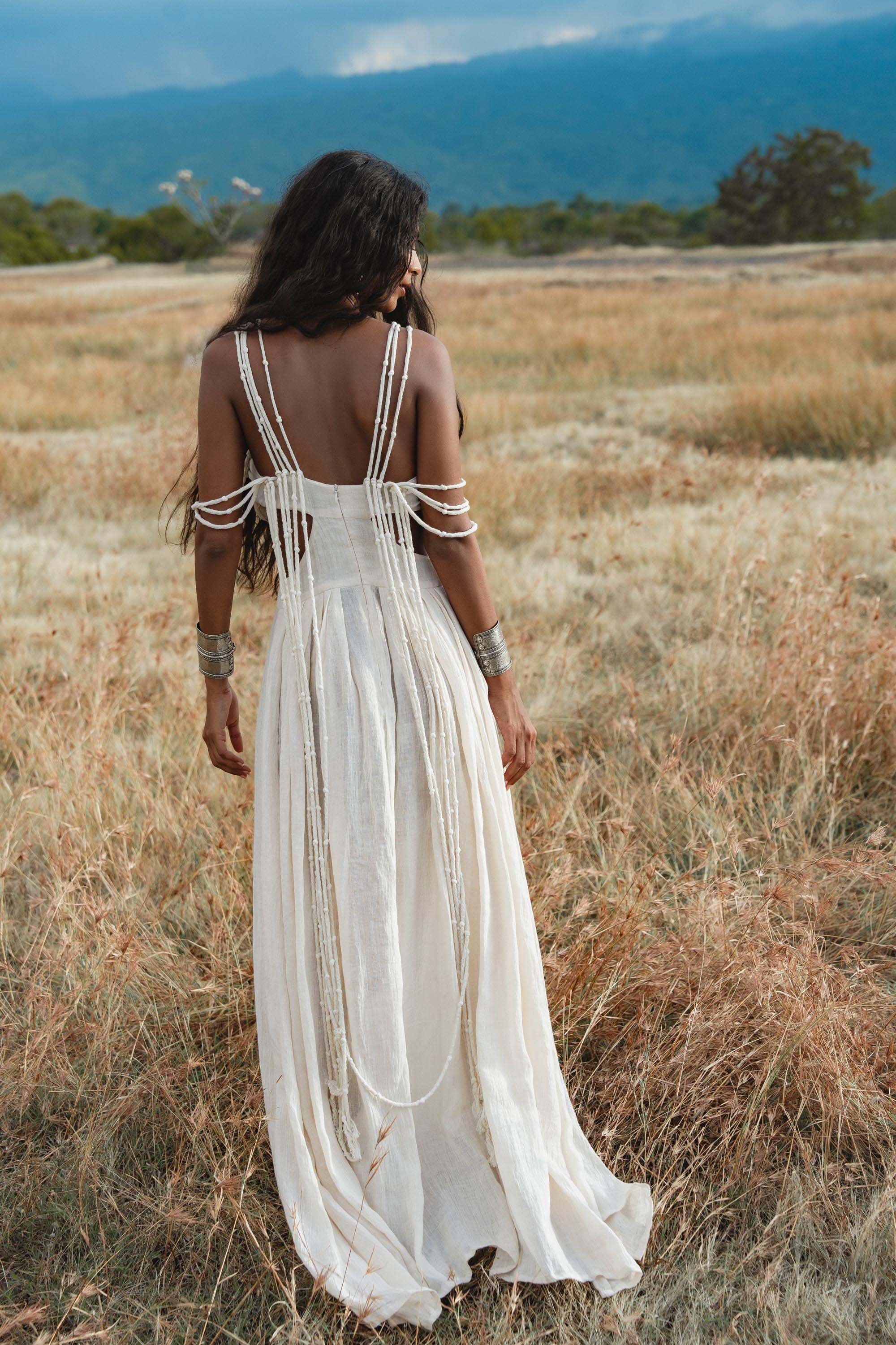 A woman with long wavy dark hair stands in a dry grassy field, facing away. She wears the Ananda Lālita Dress in ivory linen by AYA Sacred Wear, featuring beaded drapes over her arms and back, paired with brass rings and silver bracelets.