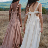 Two women stroll through a dry grassy field toward distant hills, one in the AYA Sacred Wear Ananda Lālita Dress in Ivory, its handcrafted brass rings on the straps catching the light, the other in pale blush. Flowing backless designs move with the breeze under a cloudy sky.