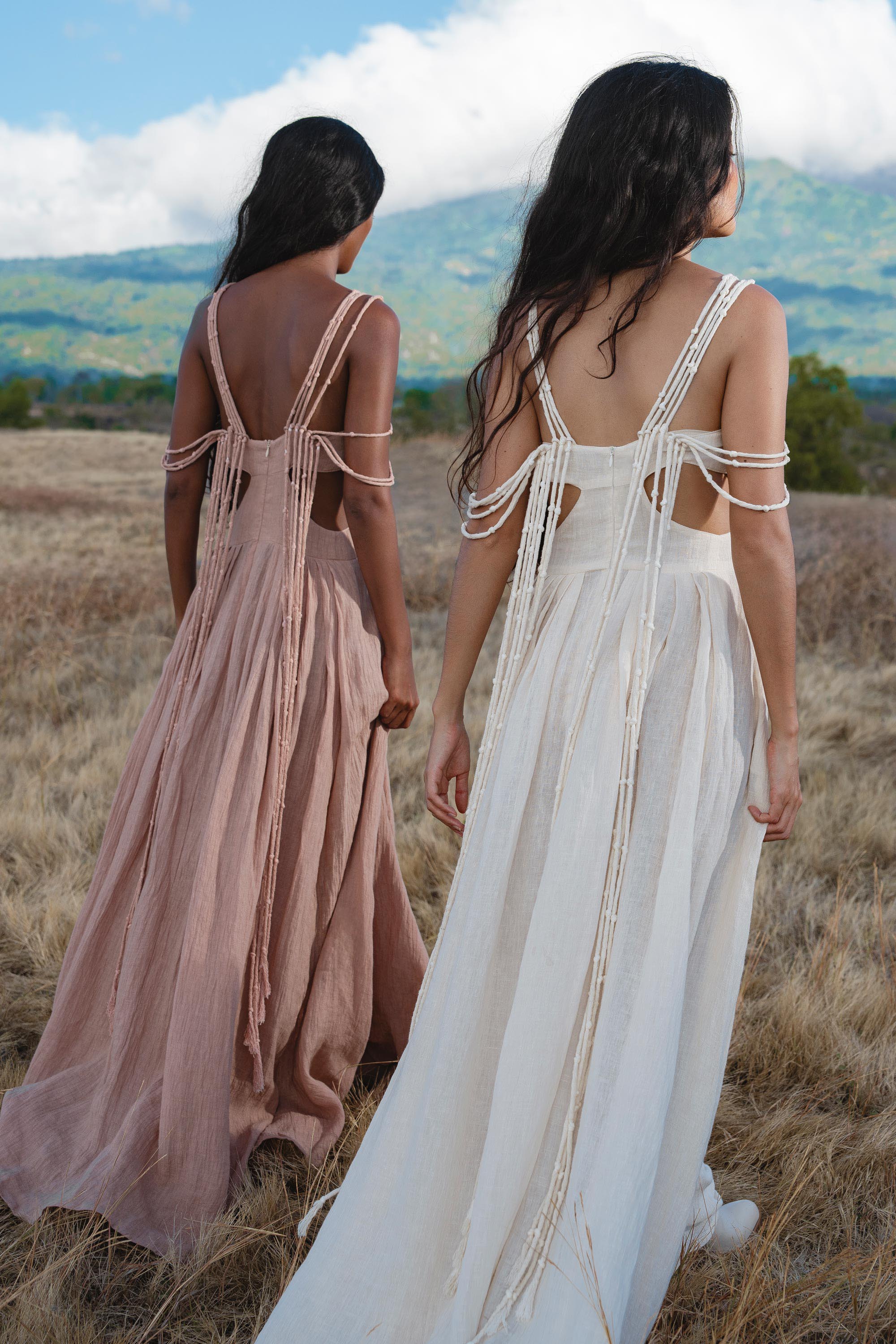 Two women stroll through a dry grassy field toward distant hills, one in the AYA Sacred Wear Ananda Lālita Dress in Ivory, its handcrafted brass rings on the straps catching the light, the other in pale blush. Flowing backless designs move with the breeze under a cloudy sky.