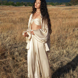 A woman with long wavy hair stands in tall grass, eyes closed and relaxed, wearing the Apsara Bra in Cream Colour – Pure Silk Transformable by AYA Sacred Wear. Green mountains and a cloudy sky form the background.