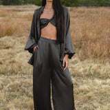 A woman with long, straight hair stands in a dry field, calmly wearing the Apsara Bra in Asphalt Gray – Pure Silk Transformable by AYA Sacred Wear, paired with loose black wide-leg pants and an open shirt. Mountains and trees are visible in the background.