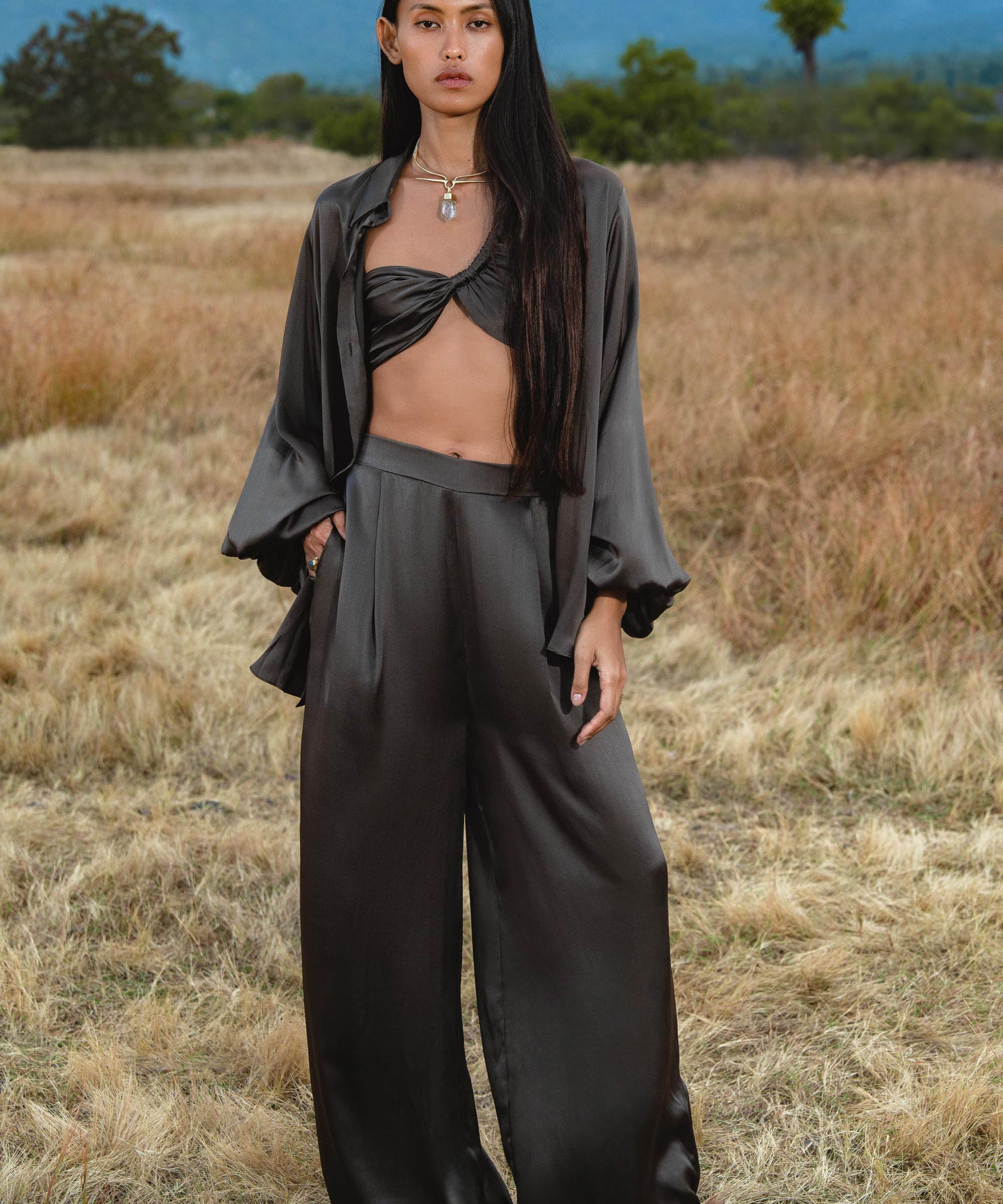A woman with long, straight hair stands in a dry field, calmly wearing the Apsara Bra in Asphalt Gray – Pure Silk Transformable by AYA Sacred Wear, paired with loose black wide-leg pants and an open shirt. Mountains and trees are visible in the background.
