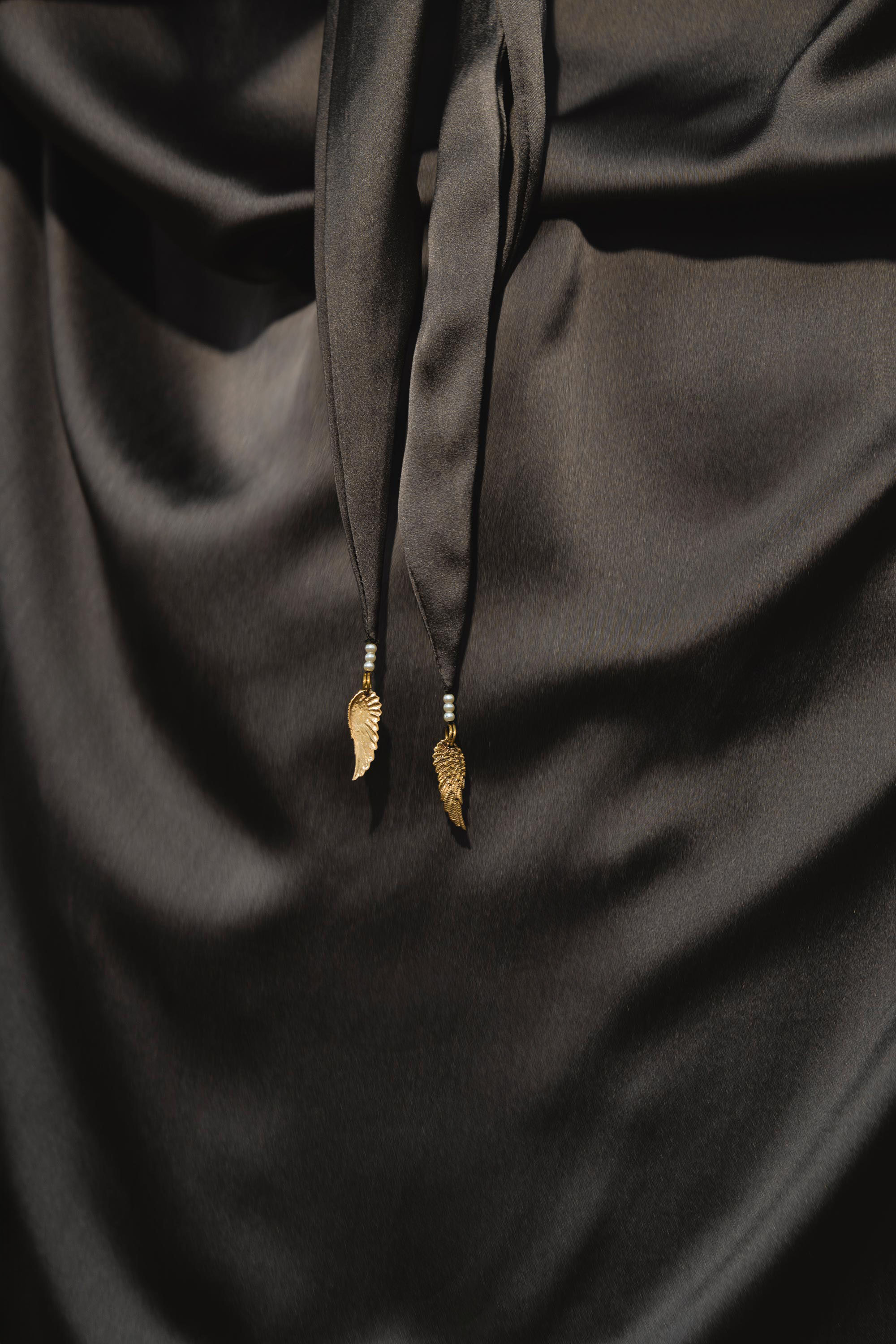 A close-up of the AYA Sacred Wear Apsara Bra in Asphalt Gray – Pure Silk Transformable shows smooth, shiny silk fabric and two black strings tied in a knot, each finished with a handcrafted gold wing charm for elegant detail.