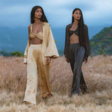 Two women walk side by side in a dry field with mountains behind them. One wears the Apsara Bra in Asphalt Gray by AYA Sacred Wear—pure silk, transformable, and handcrafted in Bali for effortless style and artistry.
