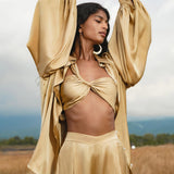 A woman stands in a field with arms raised, wearing the AYA Sacred Wear Apsara Bra in Inka Gold—pure silk, transformable. She has long dark hair, large hoop earrings, and is outdoors under a cloudy sky with distant hills.