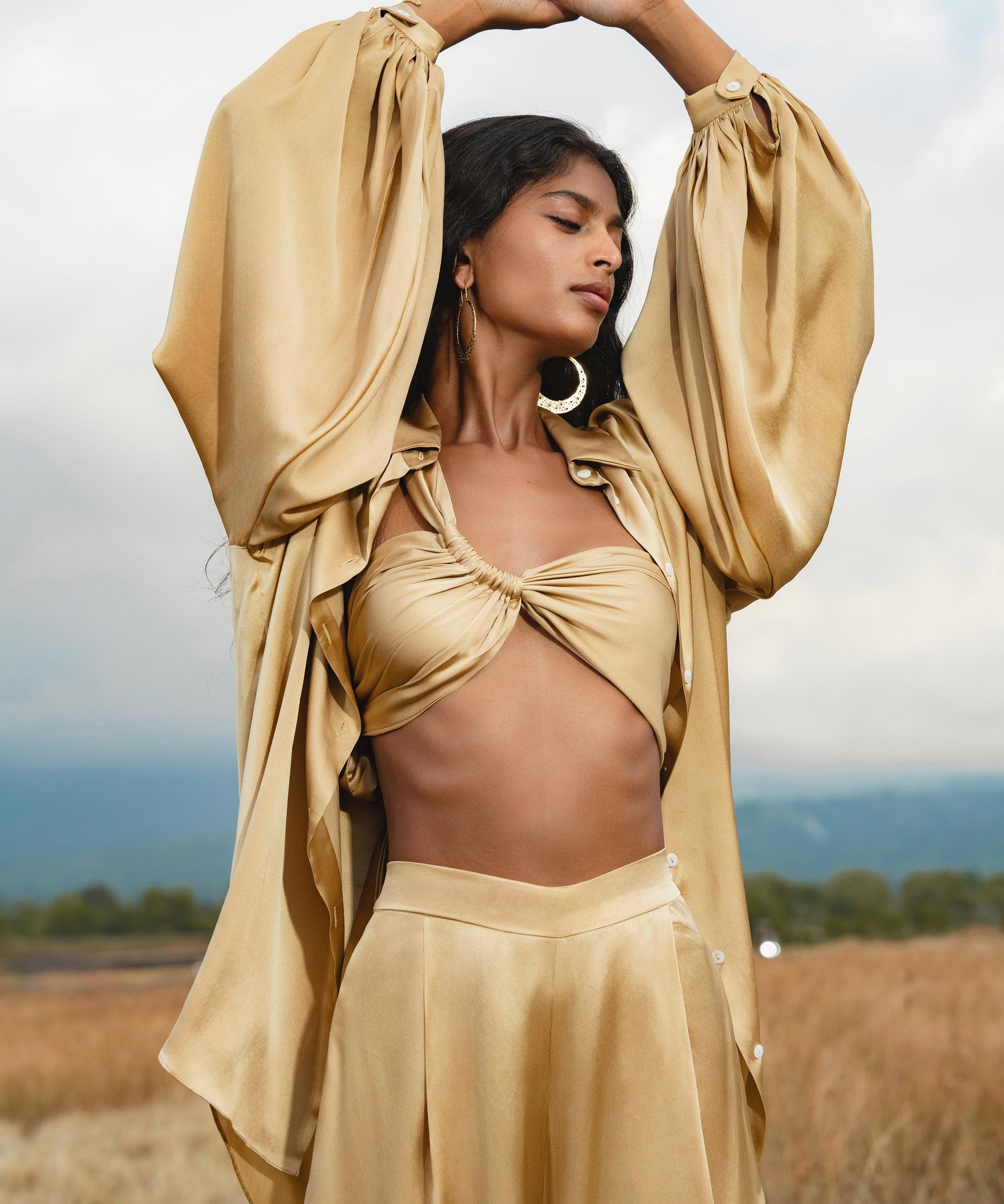 A woman stands in a field with arms raised, wearing the AYA Sacred Wear Apsara Bra in Inka Gold—pure silk, transformable. She has long dark hair, large hoop earrings, and is outdoors under a cloudy sky with distant hills.