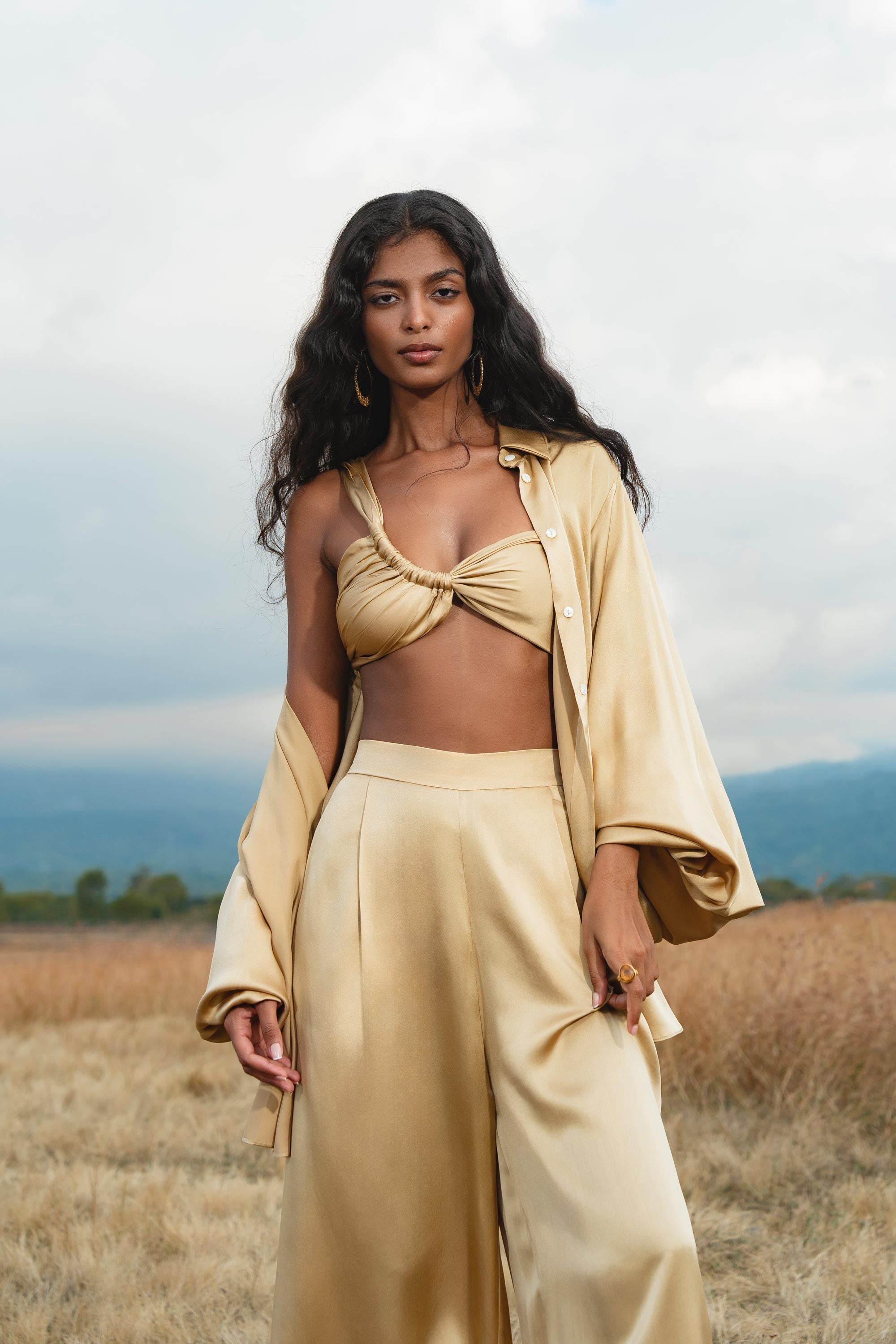 A woman with long, wavy dark hair stands in a dry grassy field, wearing the Apsara Bra in Inka Gold – Pure Silk Transformable by AYA Sacred Wear. Cloudy skies and hills form the background.