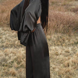 A woman with long dark hair stands in a dry grassy field, wearing loose black satin pants and the AYA Sacred Wear Apsara Kimono Blouse in Asphalt Gray—a pure silk kimono handcrafted in Bali. She gazes downward, hills softly blurred behind her.