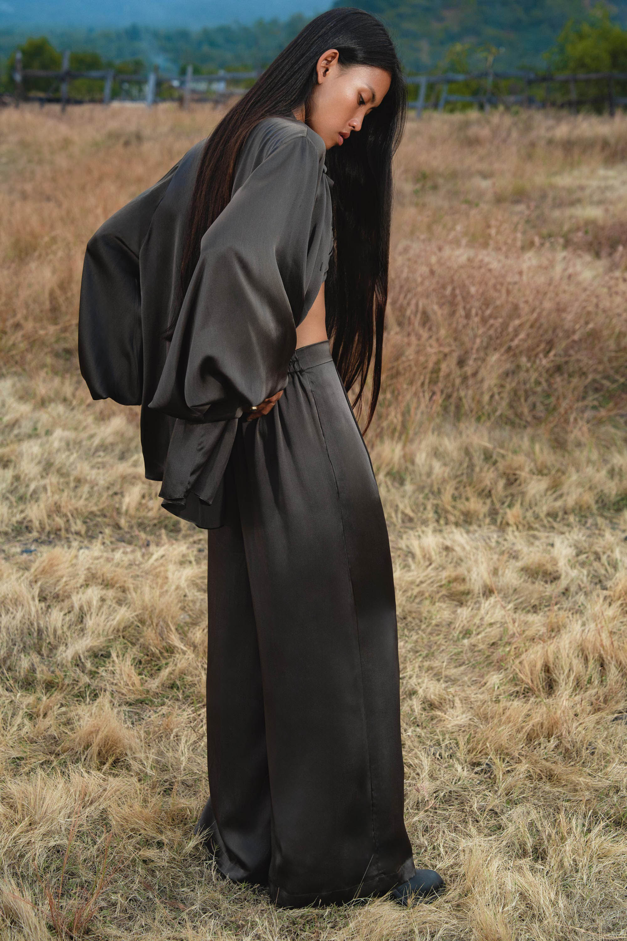 A woman with long dark hair stands in a dry grassy field, wearing loose black satin pants and the AYA Sacred Wear Apsara Kimono Blouse in Asphalt Gray—a pure silk kimono handcrafted in Bali. She gazes downward, hills softly blurred behind her.