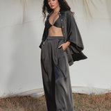 A woman stands on dry grass before a cream fabric backdrop, wearing the AYA Sacred Wear Apsara Kimono Blouse in Asphalt Gray—a pure silk kimono—over a black bralette and wide-legged pants. She looks confidently at the camera under a partly cloudy sky.
