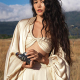 A woman with long, wavy dark hair stands under a blue sky, confidently holding a vintage camera. She wears the AYA Sacred Wear Apsara Kimono Blouse Cream – Pure Silk Kimono, its satin and puffy sleeves glowing against the mountains.