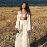 A woman with long wavy hair stands in a dry grassy field, wearing the AYA Sacred Wear Apsara Kimono Blouse Cream – a pure silk kimono handcrafted in Bali. She pairs it with wide-legged pants and a bralette, gazing serenely at distant mountains.