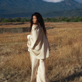 A woman with long curly hair wears the AYA Sacred Wear Apsara Kimono Blouse Cream – Pure Silk Kimono, handcrafted in Bali, standing in a dry field with green trees and blue mountains behind her beneath a partly cloudy sky.