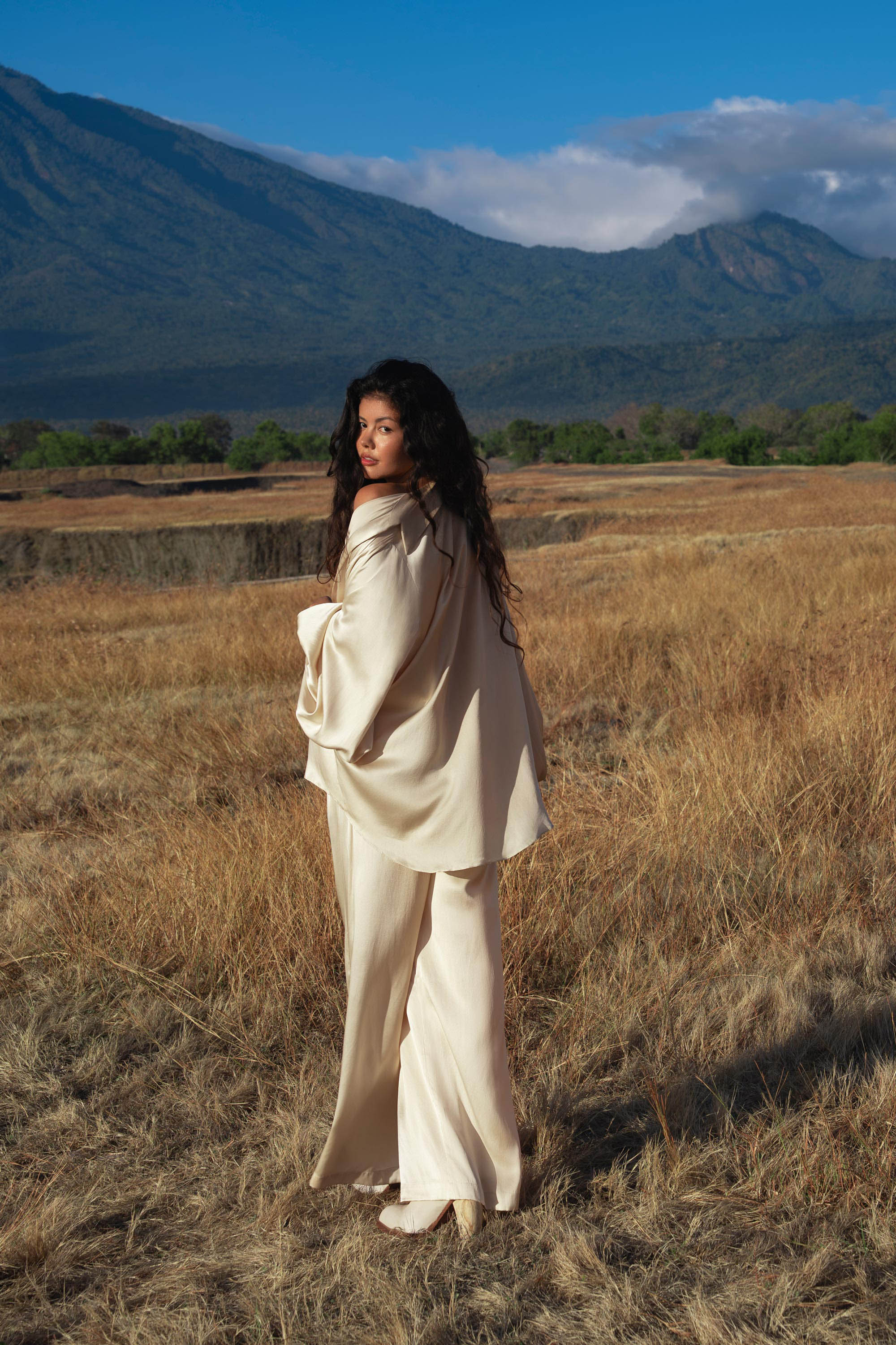A woman with long curly hair wears the AYA Sacred Wear Apsara Kimono Blouse Cream – Pure Silk Kimono, handcrafted in Bali, standing in a dry field with green trees and blue mountains behind her beneath a partly cloudy sky.