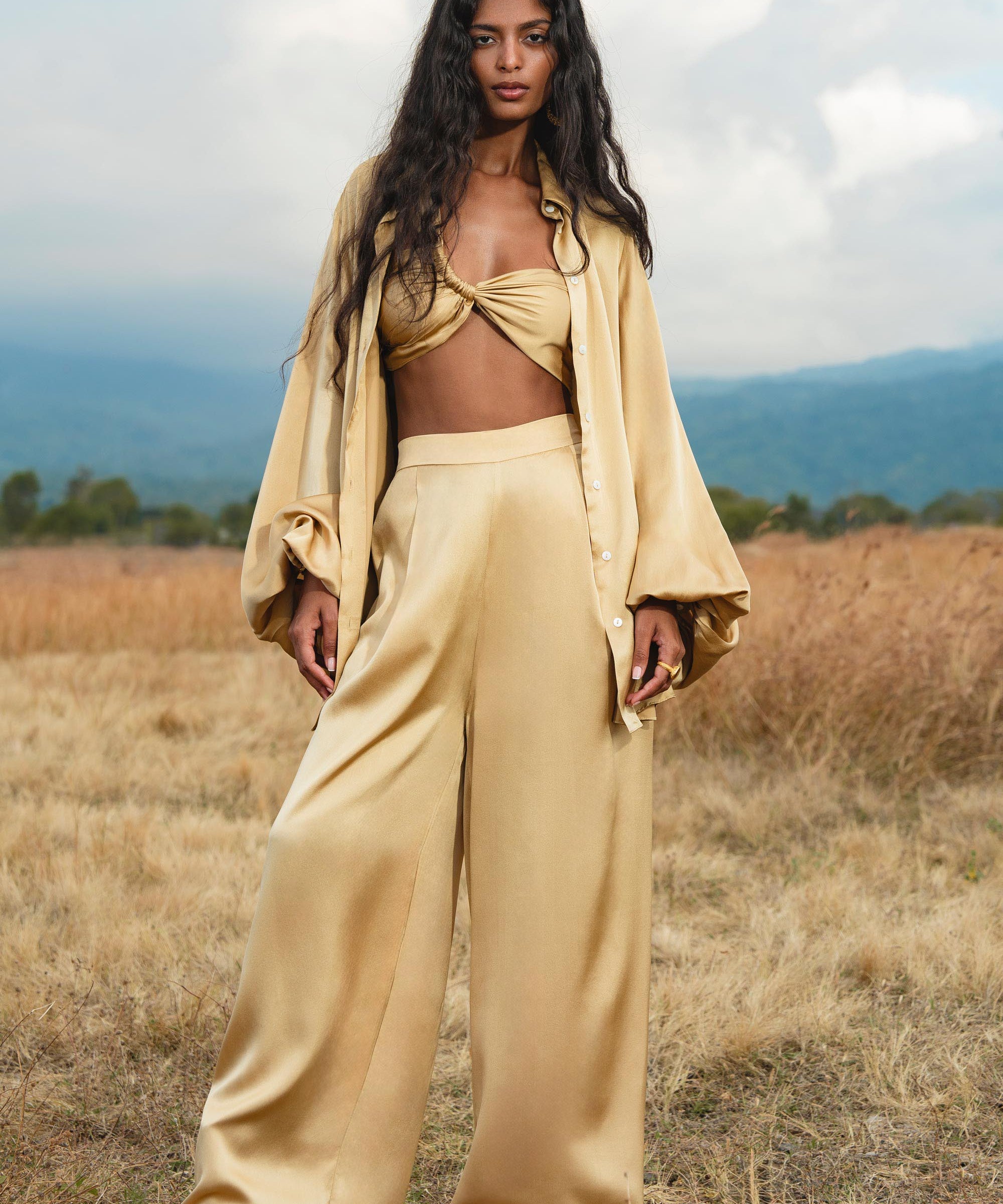 A woman with long, wavy black hair stands in a dry grassy field, wearing the AYA Sacred Wear Apsara Kimono Blouse Inka Gold—an elegant pure silk top for women—as wide-leg trousers complete her chic look amid hills and a cloudy sky.