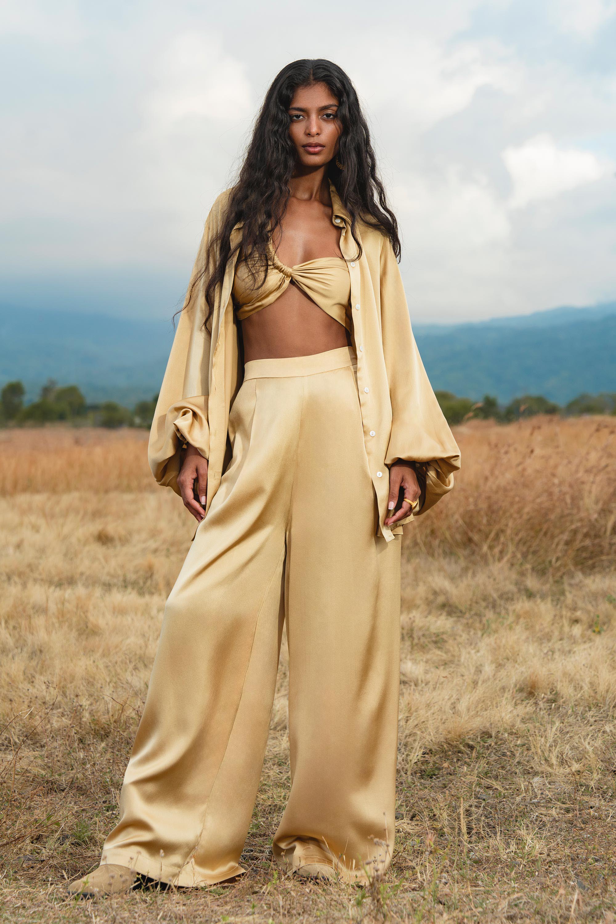 A woman with long, wavy black hair stands in a dry grassy field, wearing the AYA Sacred Wear Apsara Kimono Blouse Inka Gold—an elegant pure silk top for women—as wide-leg trousers complete her chic look amid hills and a cloudy sky.