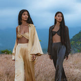 Two women stand in a grassy field with mountains behind. One wears the AYA Sacred Wear Apsara Kimono Blouse Inka Gold, handcrafted from pure silk, paired with wide pants; the other sports a dark brown bralette and matching pants for a calm, stylish look.