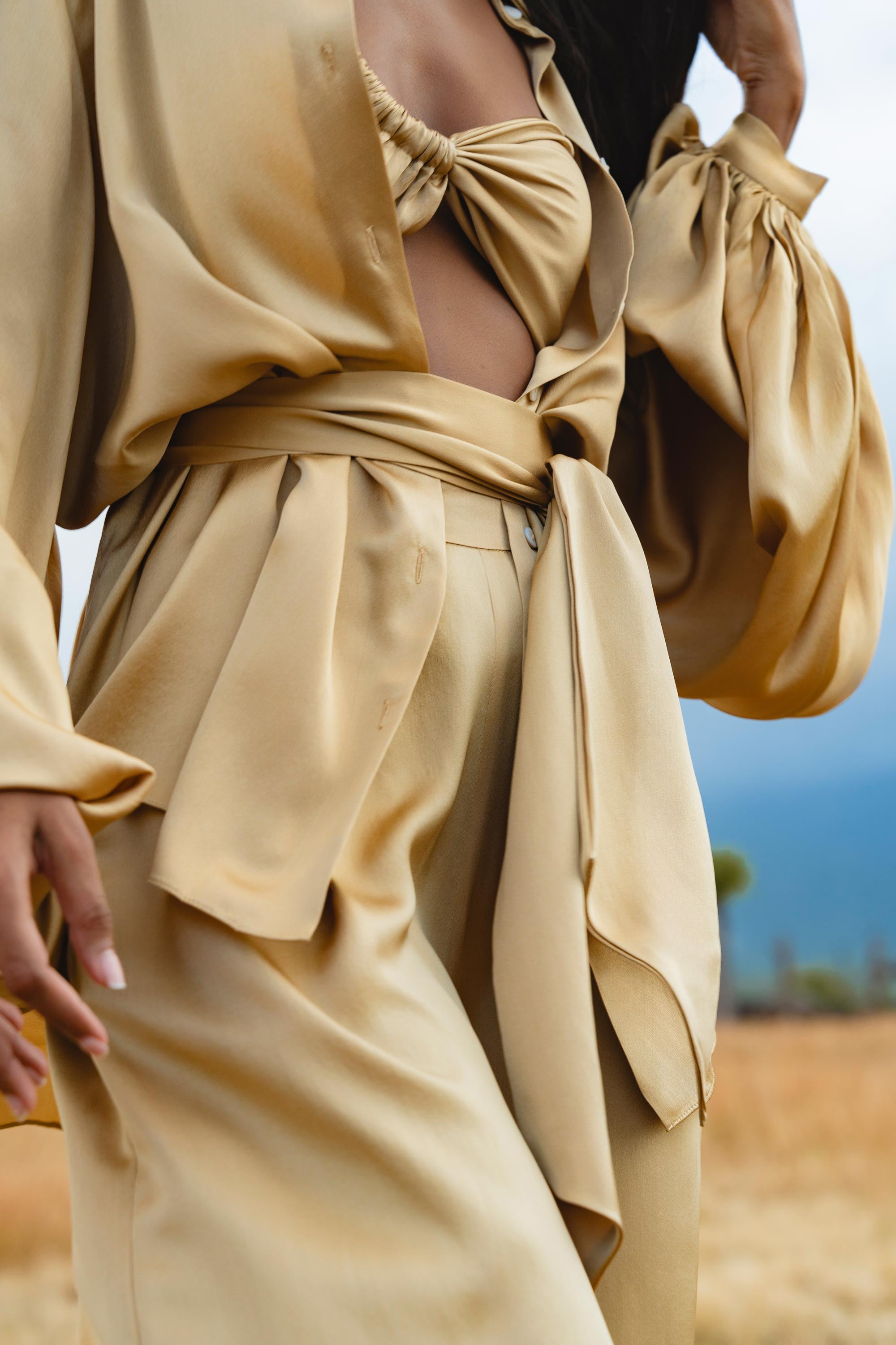 Close-up of a person wearing the Apsara Kimono Blouse Inka Gold by AYA Sacred Wear—a pure silk women's top with voluminous sleeves and a waist tie. The stylish light gold outfit stands out against a blurred outdoor scene with dry grass and blue sky.