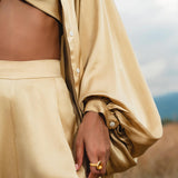 Wearing the AYA Sacred Wear Apsara Kimono Blouse Inka Gold—a pure silk top for women—paired with a bandeau and high-waisted pants, a person stands in a field, gold ring visible, with only their torso and hand shown.