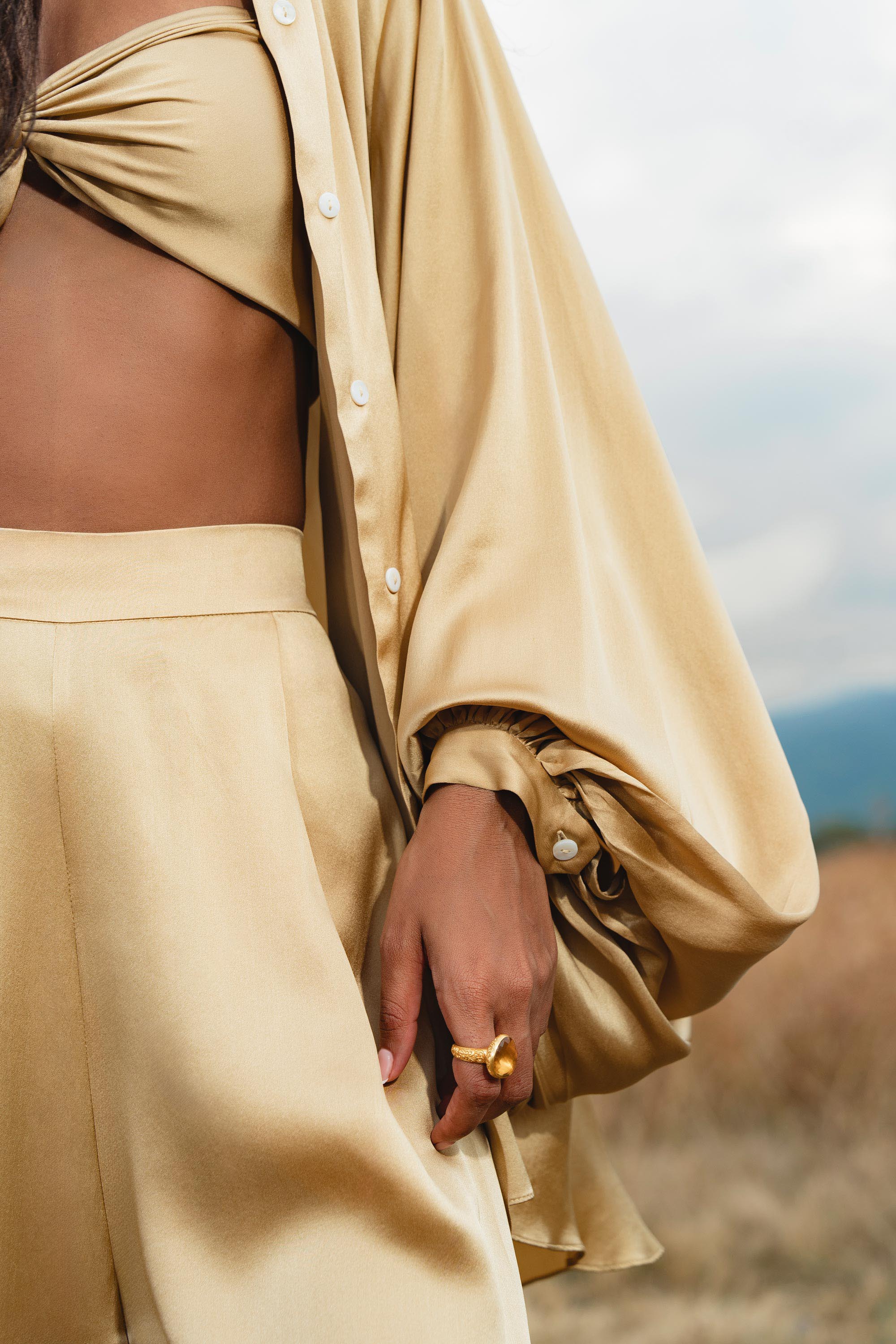 Wearing the AYA Sacred Wear Apsara Kimono Blouse Inka Gold—a pure silk top for women—paired with a bandeau and high-waisted pants, a person stands in a field, gold ring visible, with only their torso and hand shown.