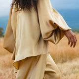 A woman with long, wavy dark hair wears the AYA Sacred Wear Apsara Kimono Blouse Inka Gold – a pure silk top for women – paired with wide pants, walking through tall golden grass under a cloudy sky with distant blue mountains. Face not visible.