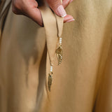 A hand holds the end of a beige Pure Silk sash adorned with gold feather charms and pearls, part of the handcrafted AYA Sacred Wear Apsara Kimono Blouse Inka Gold for women, shown on smooth, gold-toned fabric.