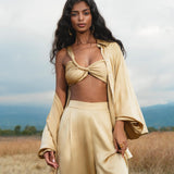 A woman with long, wavy dark hair stands in a dry field wearing the Apsara Kimono Blouse Inka Gold, a pure silk top for women by AYA Sacred Wear. She pairs it with gold hoop earrings and gazes confidently at the camera under a cloudy sky.