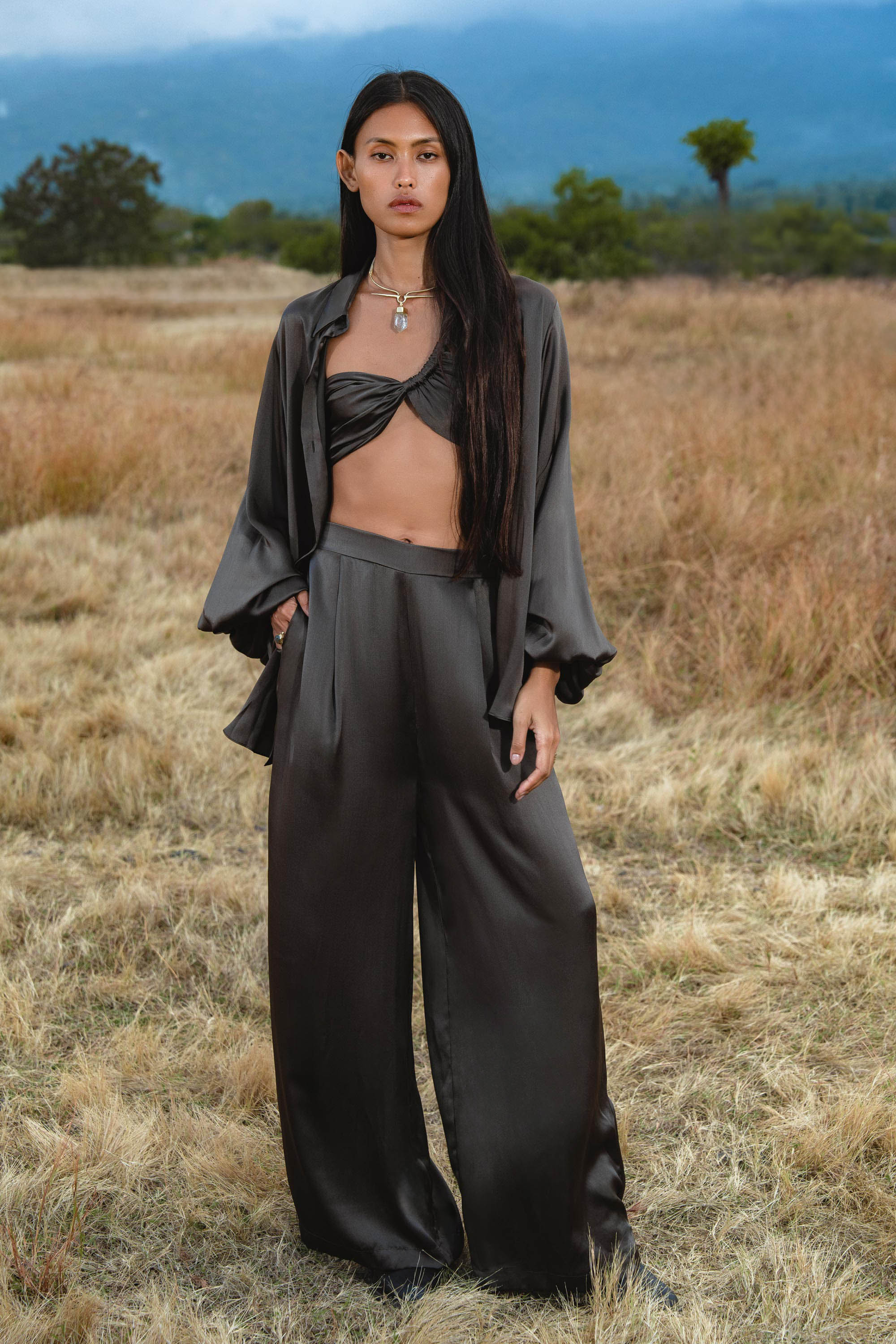 A woman with long, straight dark hair and a neutral expression stands in a dry field wearing AYA Sacred Wear’s Apsara Pants in Asphalt Gray—pure silk, flowy pants—paired with a twisted crop top and open shirt. Mountains and clouds are in the background.