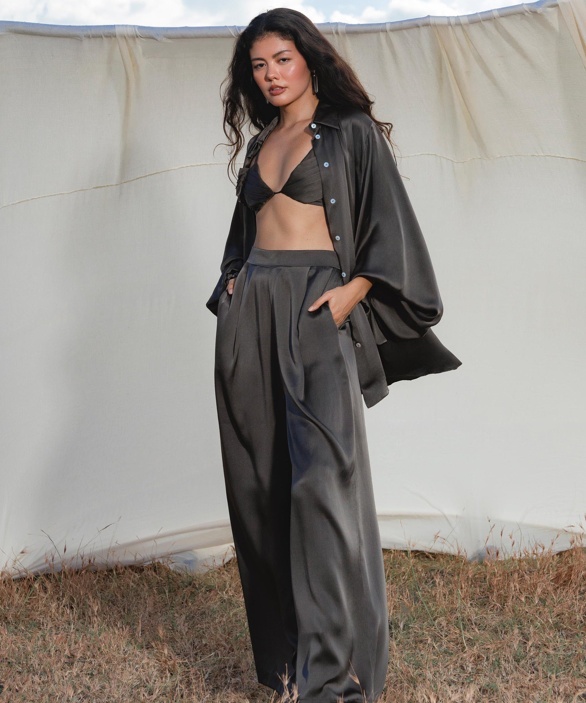 A woman with long dark hair stands on dry grass before a cream backdrop, wearing a black bikini top, oversized shirt, and AYA Sacred Wear's Apsara Pants in Asphalt Gray—pure silk flowy pants handcrafted in Bali—hands in pockets, looking confident.