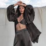 A woman poses outdoors before a cream fabric backdrop, wearing AYA Sacred Wear’s Apsara Pants in Asphalt Gray—pure silk, flowy pants—paired with a black satin bralette and an oversized black shirt, arms raised with clouds behind her.