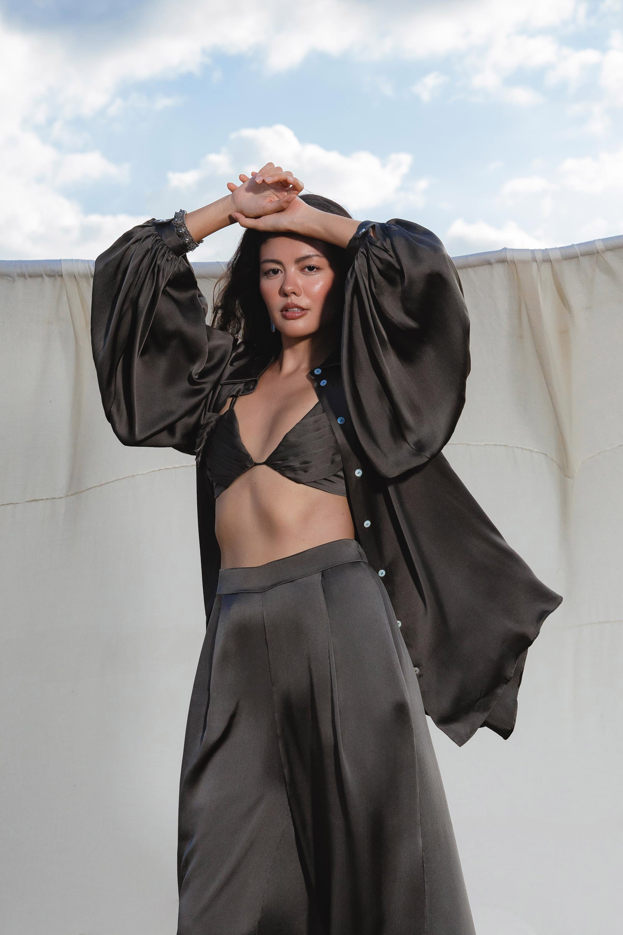 A woman poses outdoors before a cream fabric backdrop, wearing AYA Sacred Wear’s Apsara Pants in Asphalt Gray—pure silk, flowy pants—paired with a black satin bralette and an oversized black shirt, arms raised with clouds behind her.