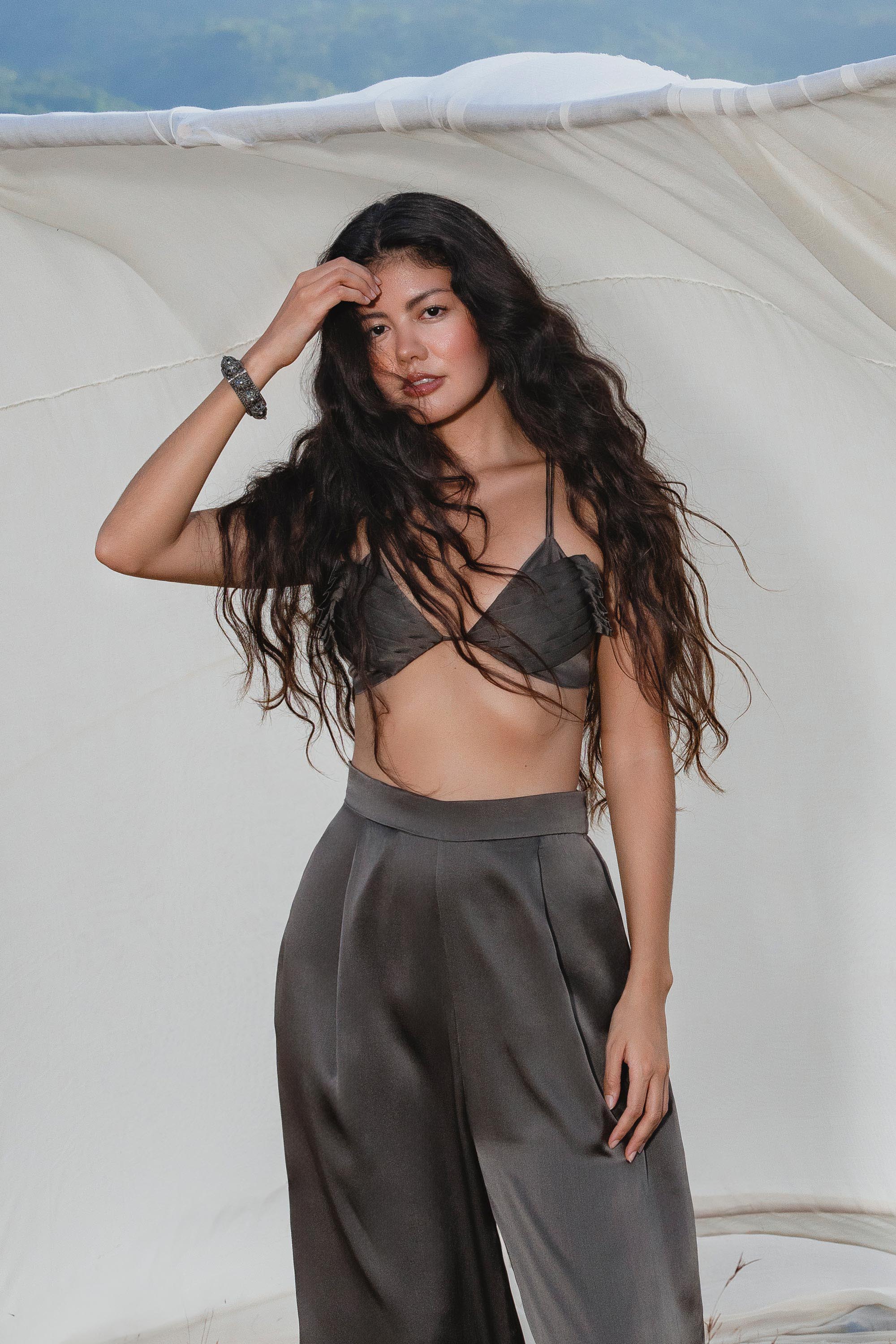 A woman with long, wavy dark hair poses outdoors by white fabric, wearing a dark bralette and AYA Sacred Wear's Apsara Pants in Asphalt Gray—pure silk flowy pants with an elegant drape—one hand in her hair, the other by her side.
