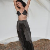 A woman stands outdoors on dry grass in front of a white sheet, wearing AYA Sacred Wear’s Apsara Pants in Asphalt Gray—pure silk, flowy, high-waisted pants—posing with arms raised and gazing at the camera under a partly cloudy sky.
