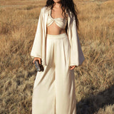 A woman stands in a dry grassy field wearing AYA Sacred Wear's Apsara Pants in cream—pure silk, flowy pants—paired with a twisted bandeau top and loose jacket. She holds a small clutch; mountains and trees are visible in the distance.