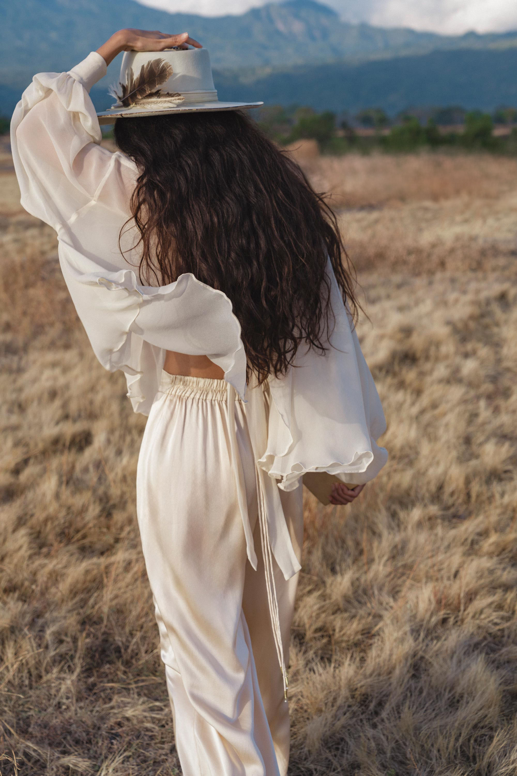 A woman with long wavy hair, dressed in AYA Sacred Wear's Apsara Pants Cream Colour – Pure Silk Flowy Pants, a cream blouse, and a decorated wide-brimmed hat, strolls through a grassy field radiating modern bohemian style.