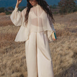 In a dry grassy field with mountain views, a woman in AYA Sacred Wear's Apsara Pants Cream Colour—pure silk flowy pants—pairs them with a sheer blouse and feathered hat. She holds a book, gazing down as her long wavy hair falls over her shoulders.
