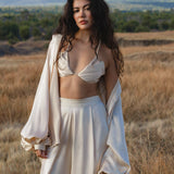 A woman with long, wavy dark hair stands in a dry field, wearing AYA Sacred Wear’s Apsara Pants in cream—pure silk, flowy elegance—with a matching oversized jacket draped over her arms. A mountain rises behind her under a partly cloudy sky.