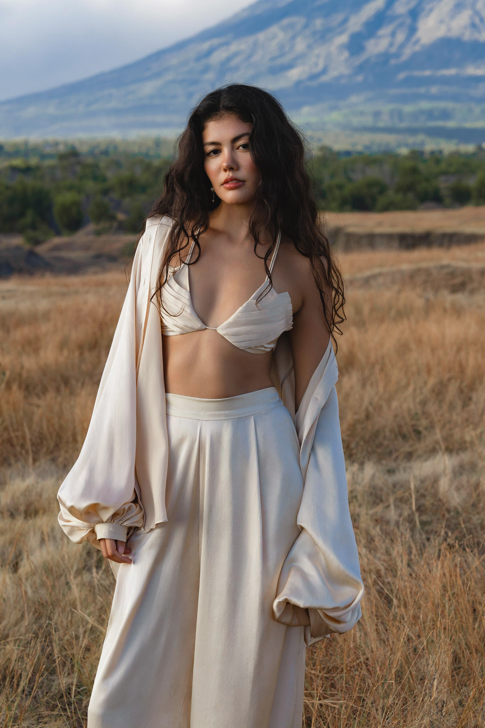 A woman with long, wavy dark hair stands in a dry field, wearing AYA Sacred Wear’s Apsara Pants in cream—pure silk, flowy elegance—with a matching oversized jacket draped over her arms. A mountain rises behind her under a partly cloudy sky.