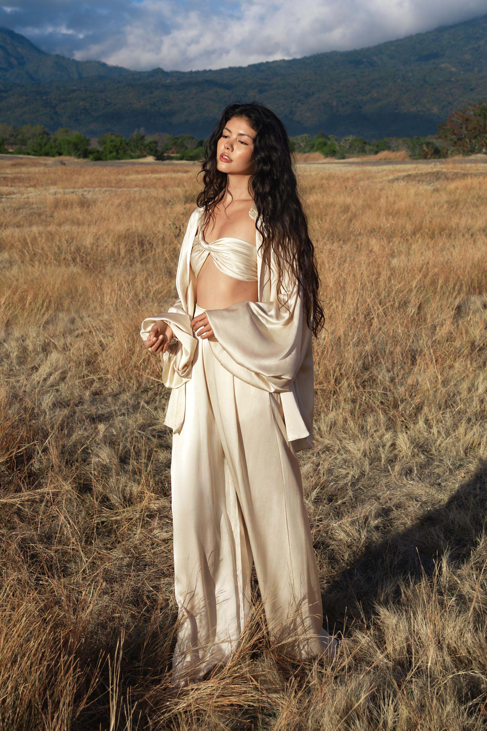 In a sunlit field, a woman in AYA Sacred Wear’s Apsara Pants—cream-coloured, pure silk, and flowy—stands with closed eyes. Her long dark hair cascades as mountains and clouds frame the serene scene.